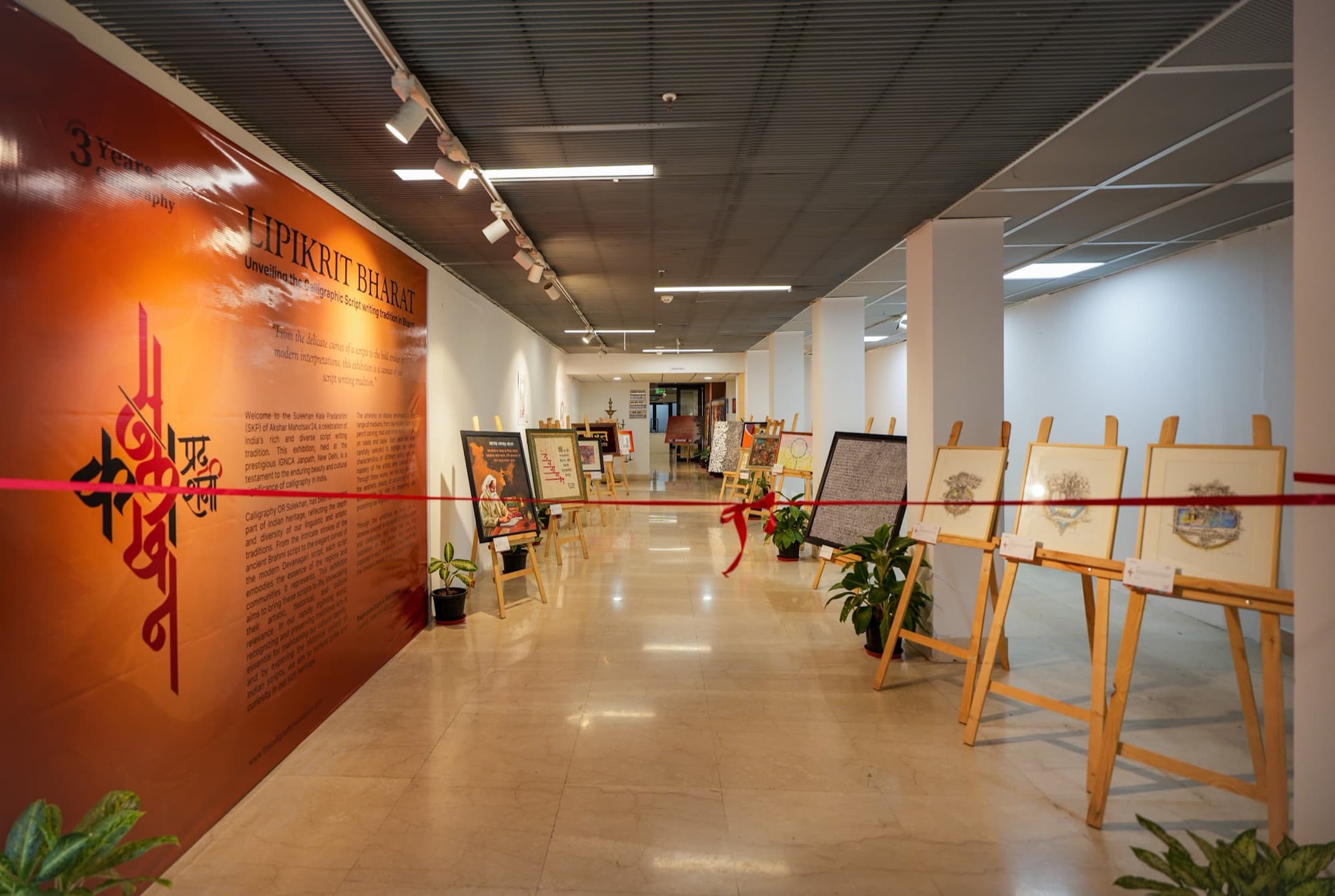 Exhibition corridor with orange calligraphy signage, works on easels and a red opening ribbon