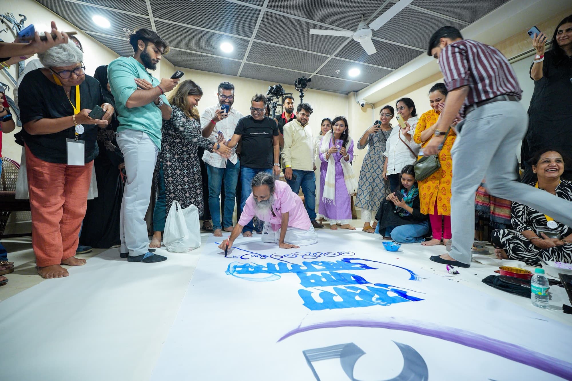 Elderly calligrapher brushing large blue letterforms on a floor canvas as a crowd photographs him