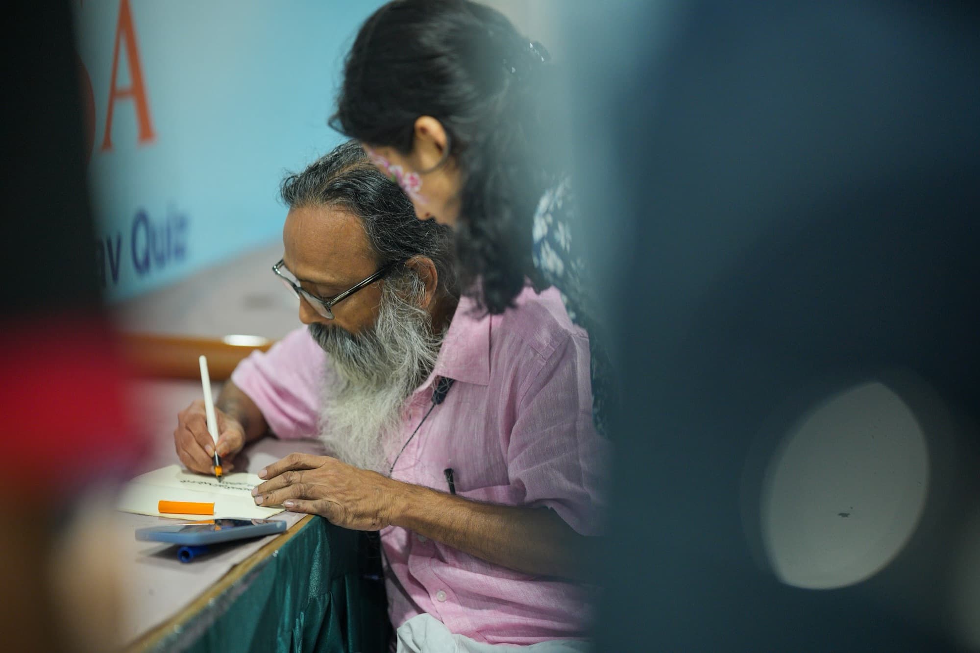 Older bearded man in glasses writing in a notebook, framed by blurred foreground figures