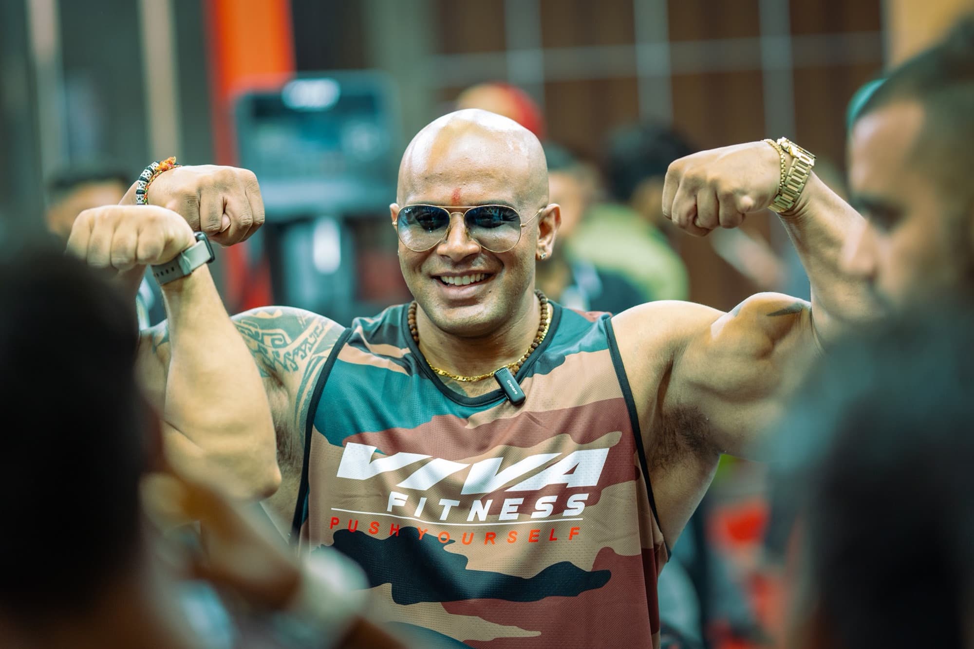 Bald bodybuilder in a camo tank hitting a double biceps pose and grinning in a crowd