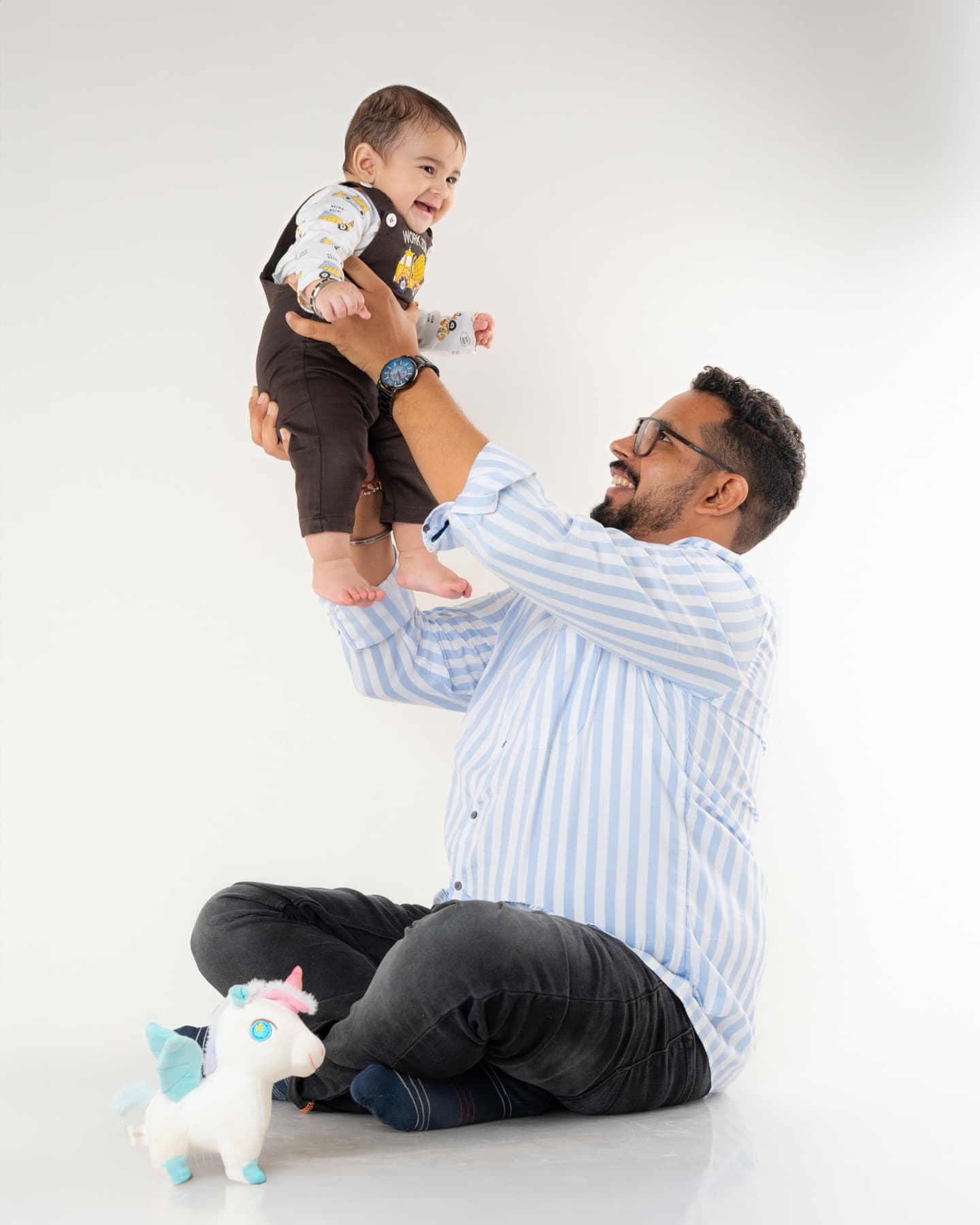 Father lifting a laughing baby overhead while seated on a white studio floor