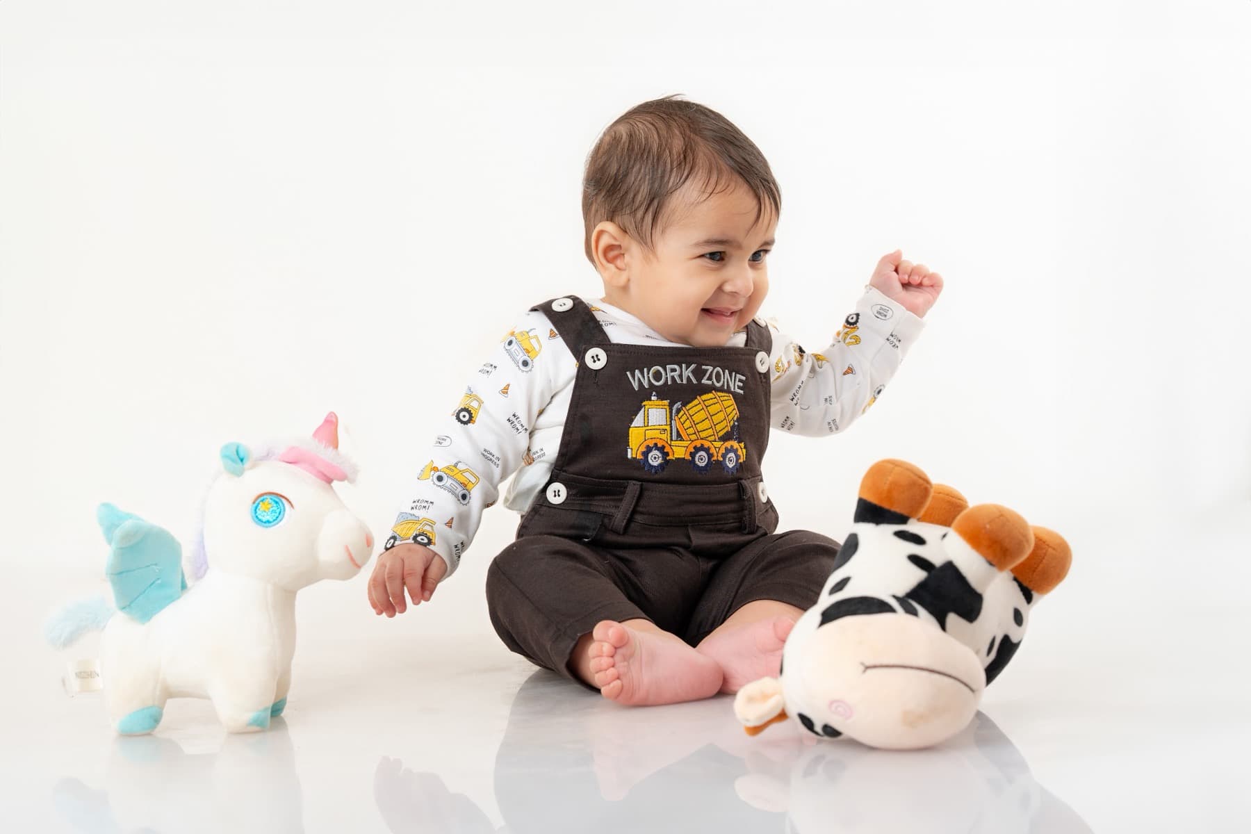 Toddler sitting on a white studio floor laughing with a fist raised, plush toys either side