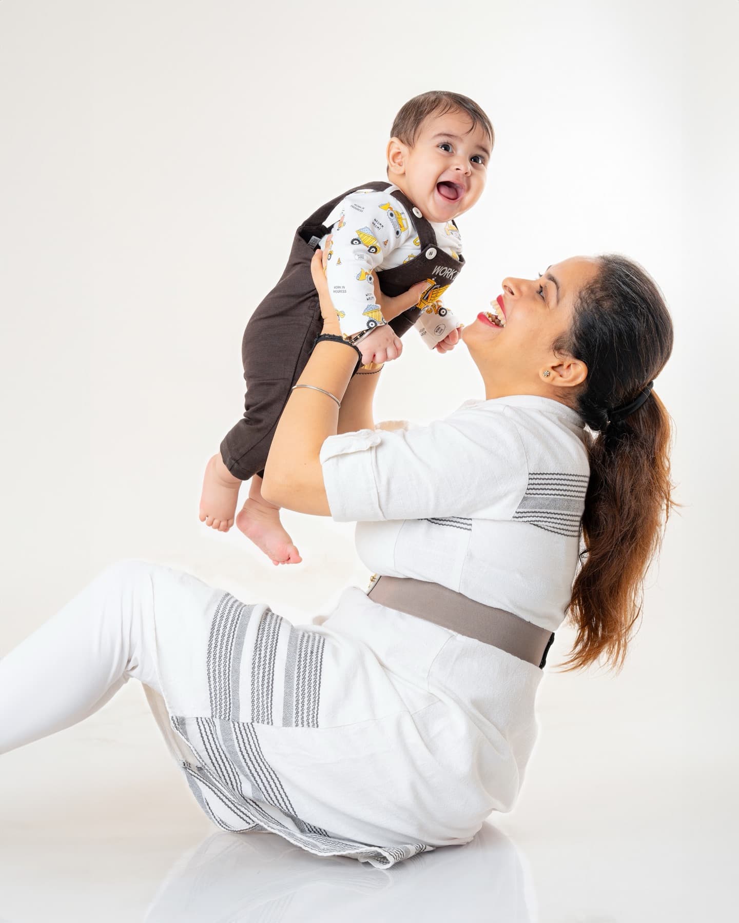 Mother seated on a studio floor lifting a laughing baby above her