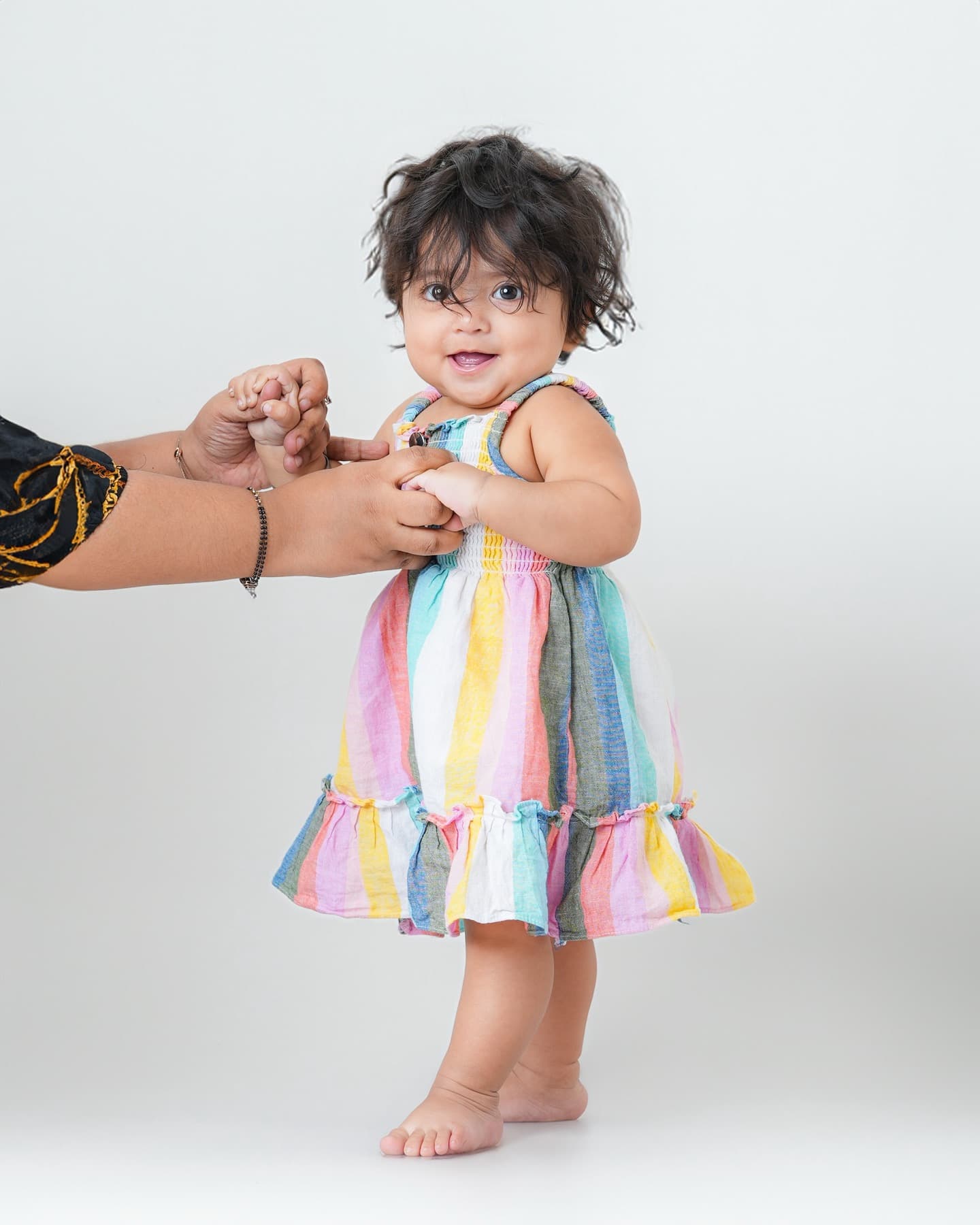 Toddler in a striped dress standing while disembodied adult hands hold her from off-frame