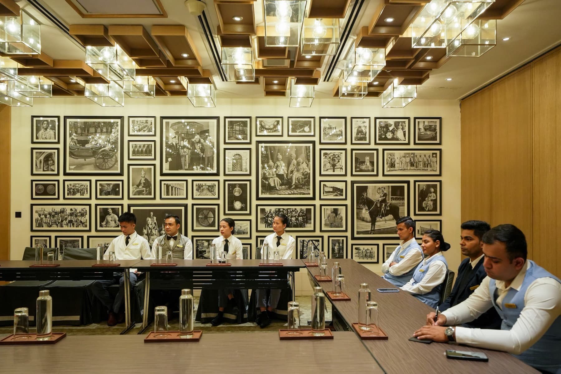 Hotel staff in waistcoats seated around a boardroom table below a wall of framed archival photos