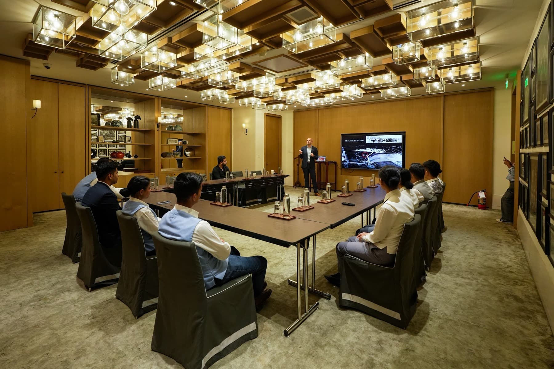 Presenter beside a screen addressing attendees around a U-shaped table under a chandelier-covered ceiling