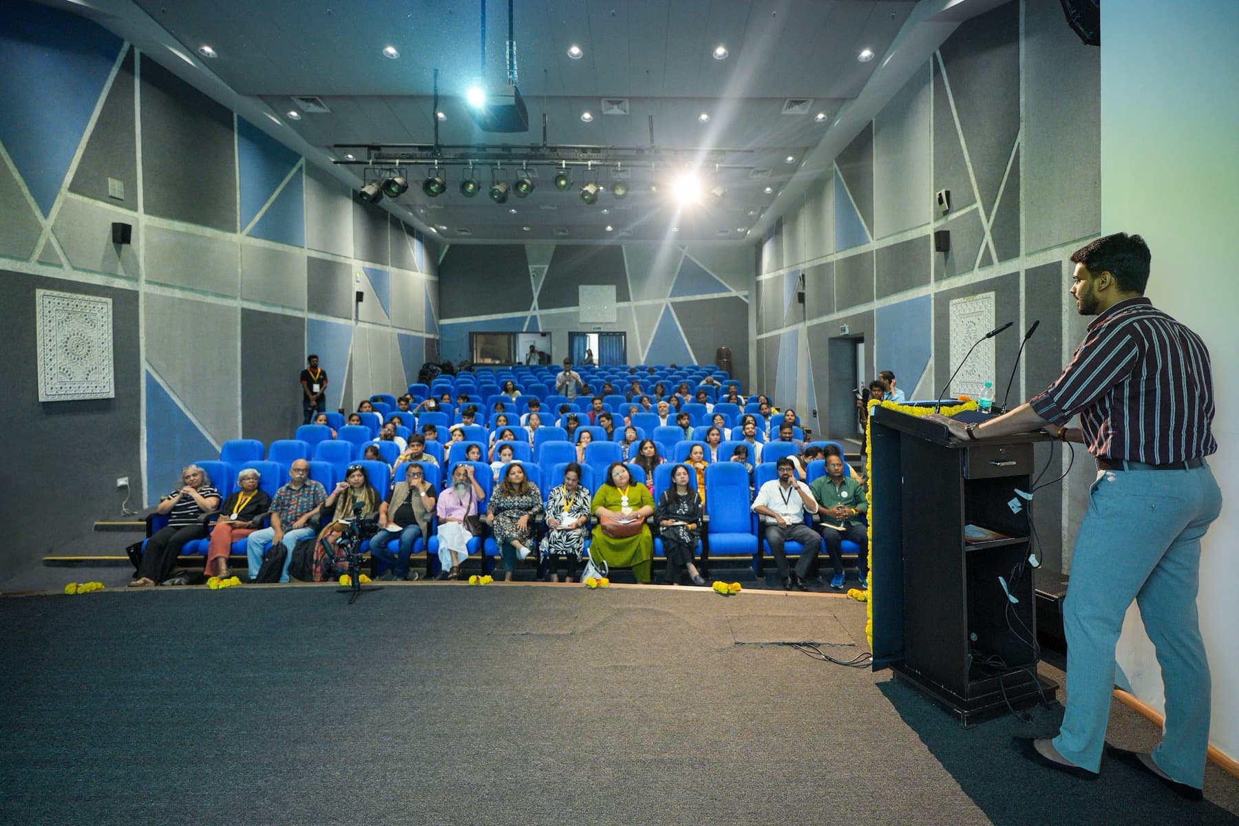 Speaker at a lectern facing an audience seated in blue theatre seats in an auditorium