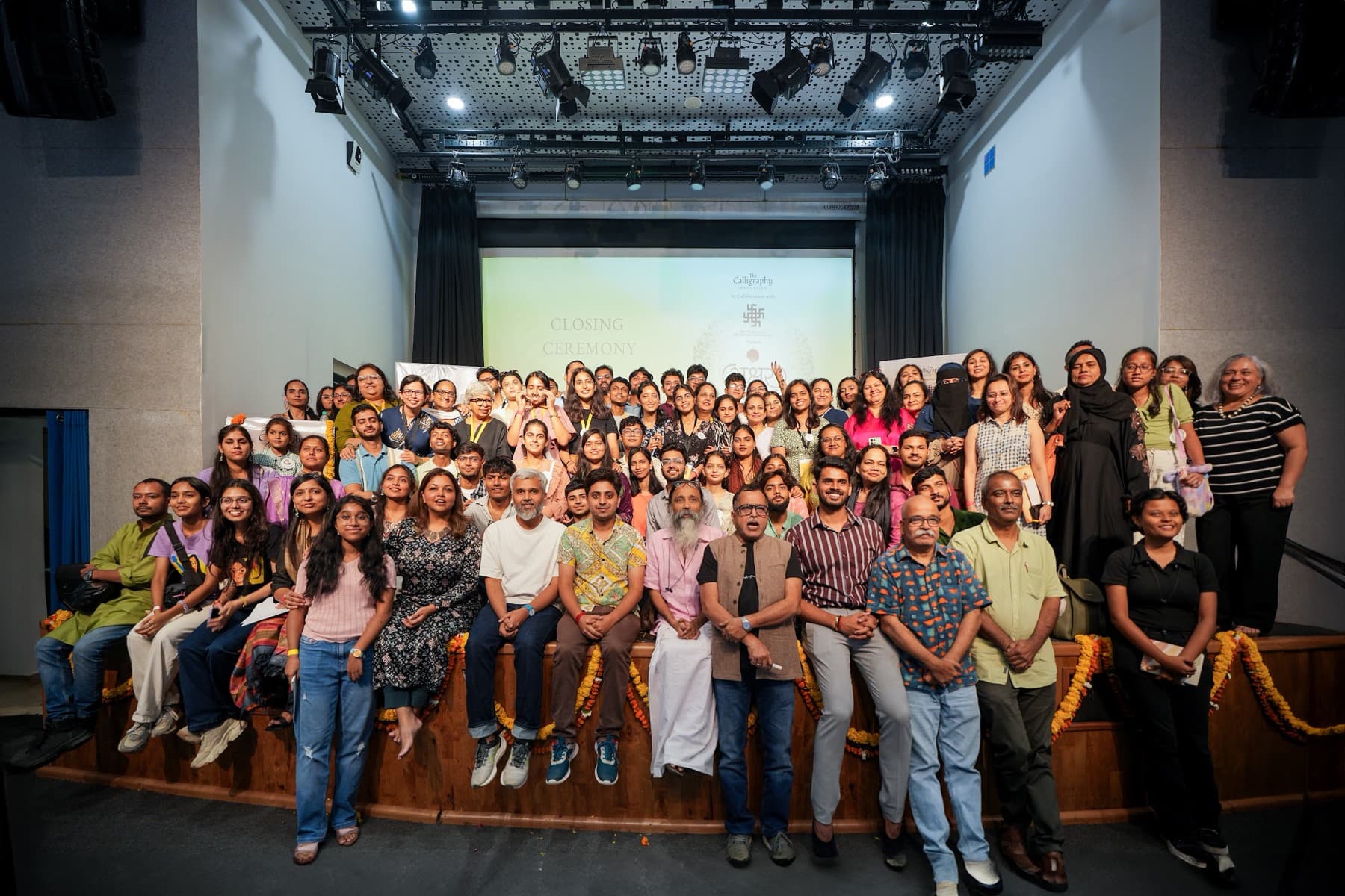 Large group photo of around eighty people on auditorium steps beneath a Closing Ceremony slide