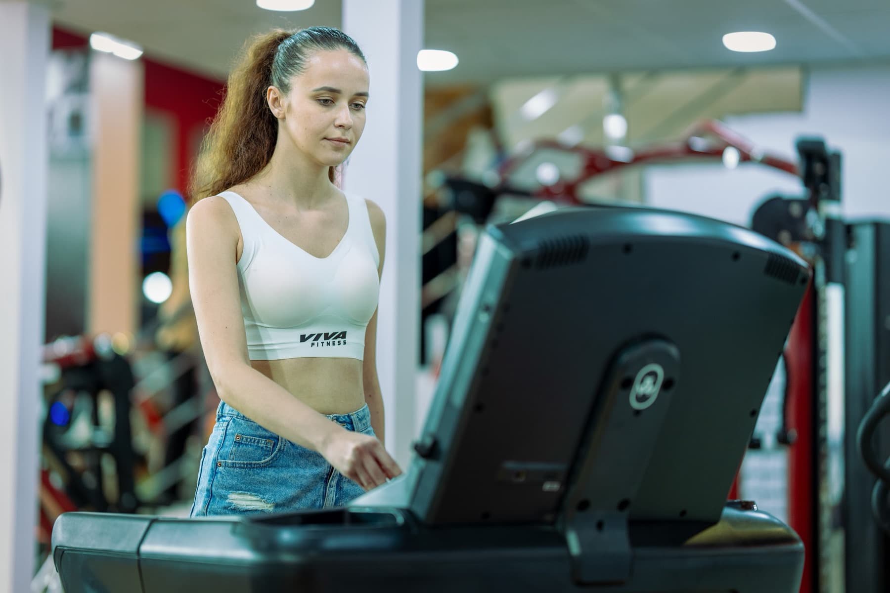 Woman in a white sports bra adjusting the console of a treadmill in a gym