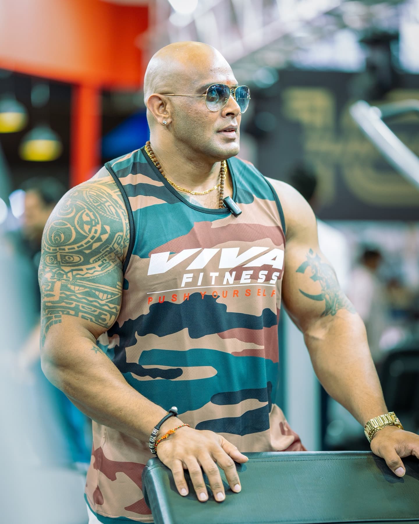 Bald bodybuilder in a camo tank leaning on gym equipment wearing sunglasses
