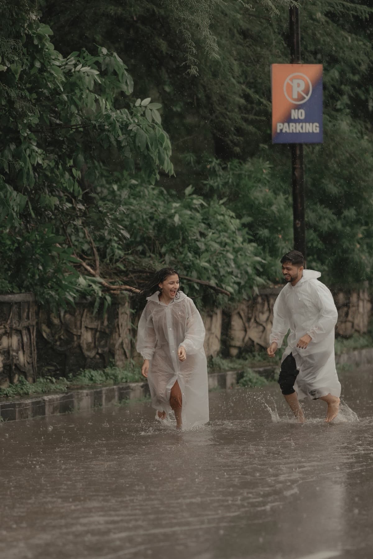 Couple in clear rain ponchos wading through a flooded road past a no-parking sign