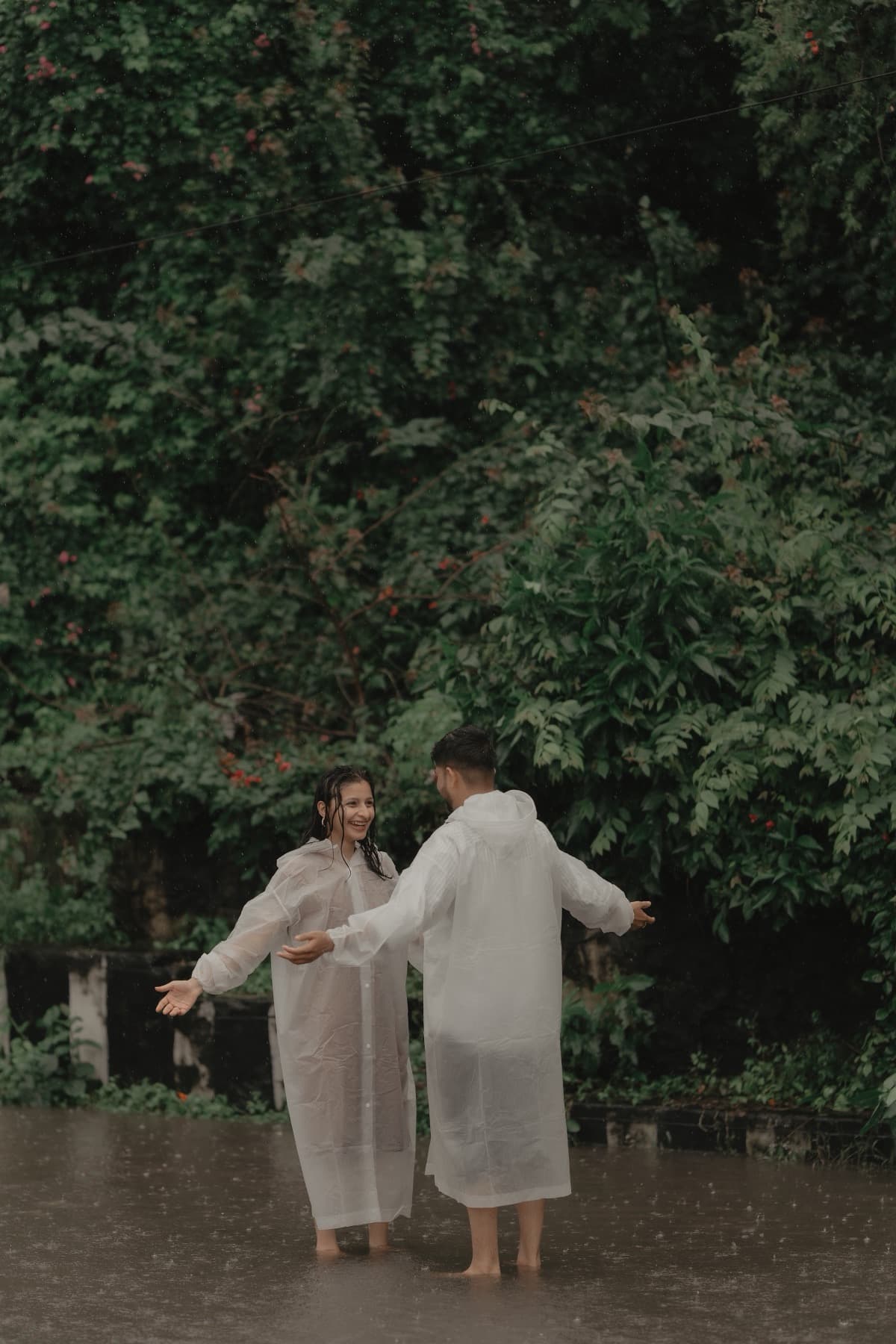 Couple in rain ponchos standing arms-out in floodwater before a green flowering bank
