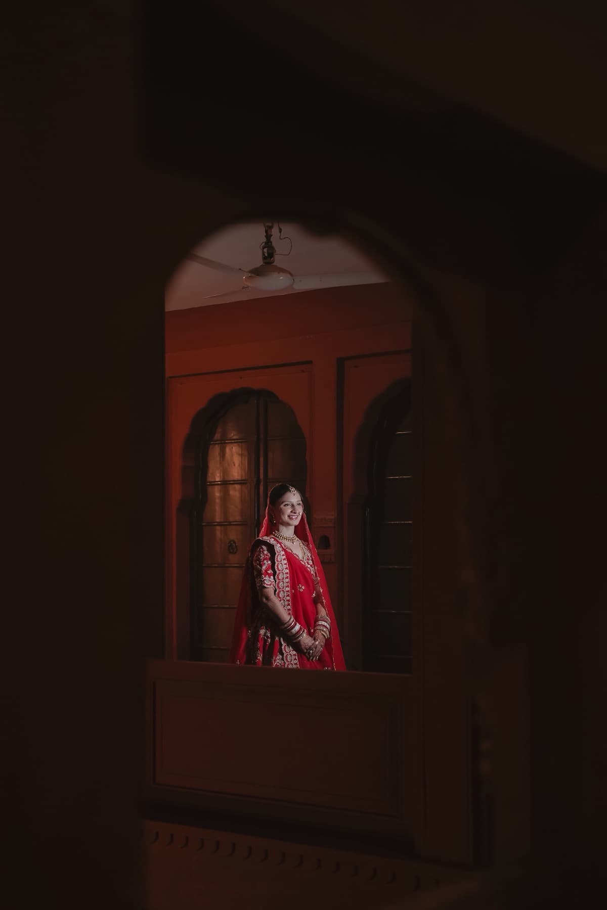Bride in a red lehenga framed in a lit doorway seen through a dark arch