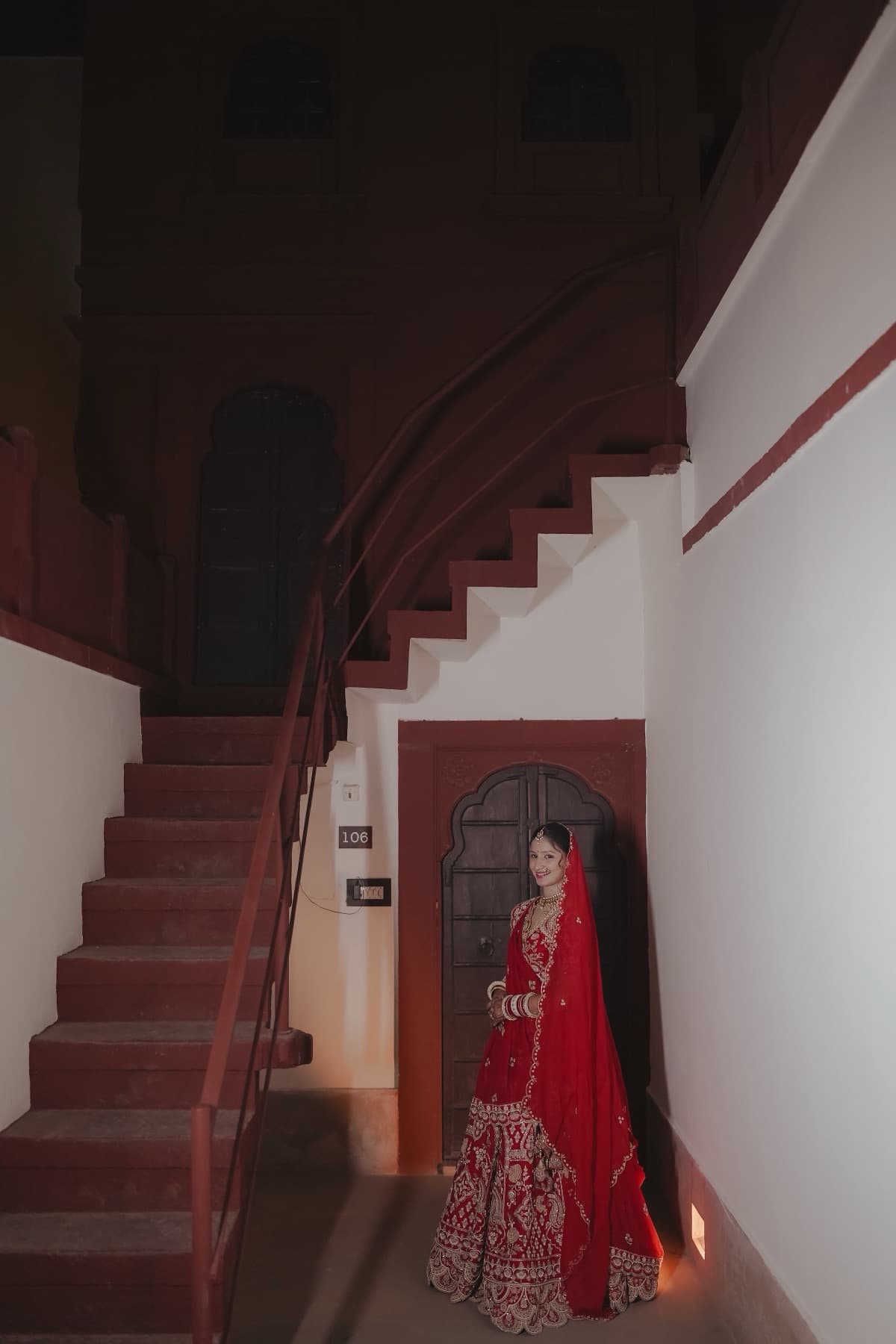 Bride in red lehenga standing beside a carved door under a curved sandstone staircase