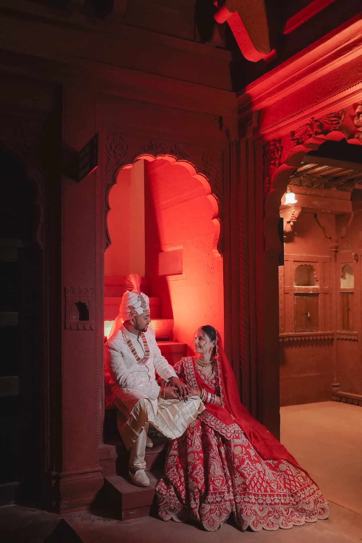 Seated couple in a red-lit carved alcove with the bride's lehenga fanned across the floor