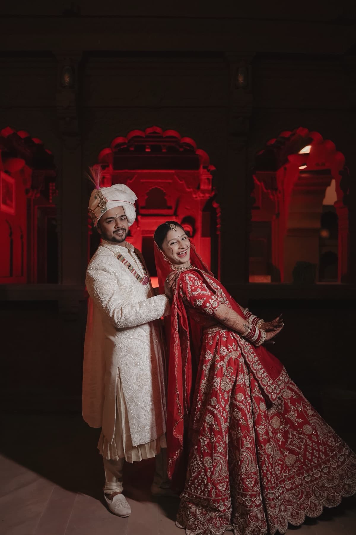 Couple smiling at camera in a colonnaded courtyard with red-lit arches behind