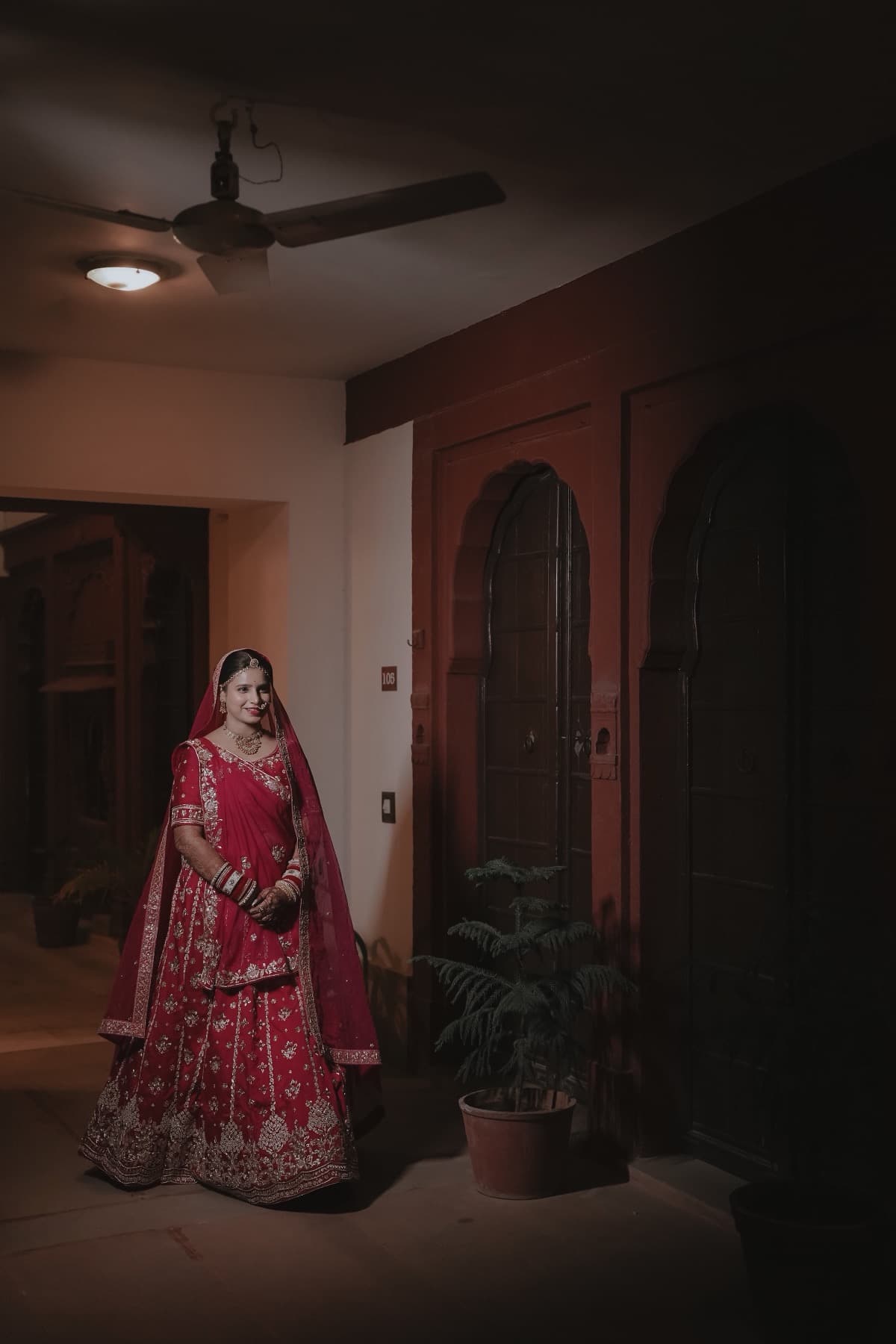 Bride in pink-red lehenga standing in a home corridor beside arched wooden doors