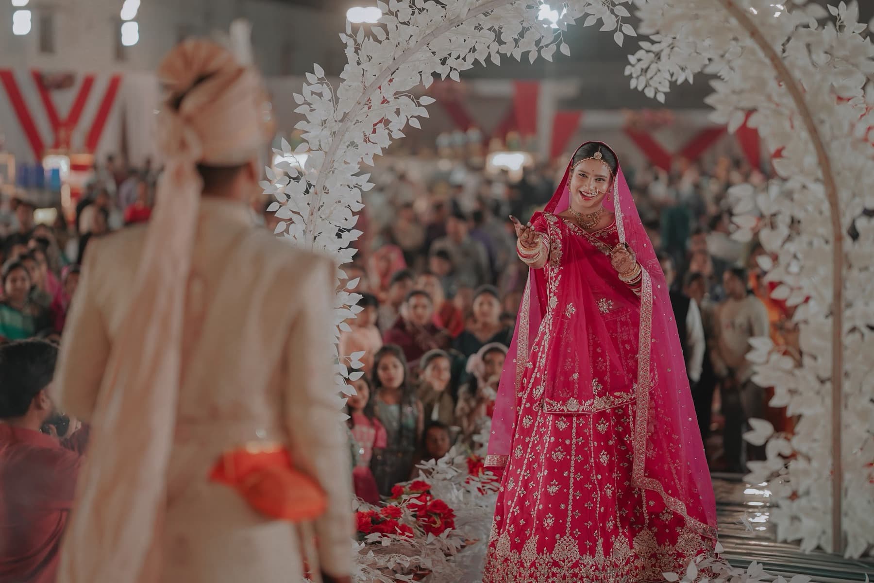 Bride walking under a white floral arch toward the groom as a hall of guests watches