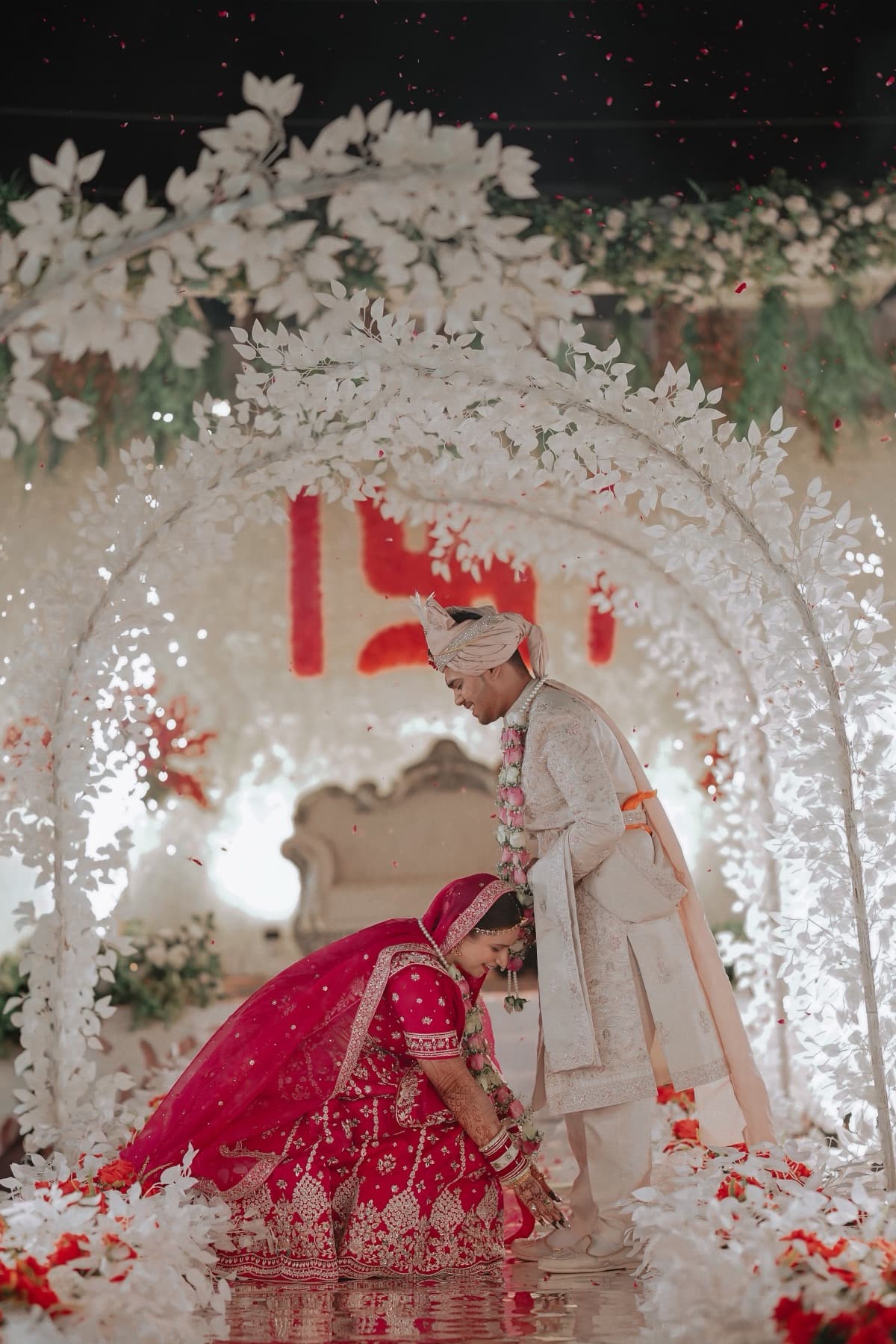 Bride bending before the standing groom on a white floral mandap strewn with petals