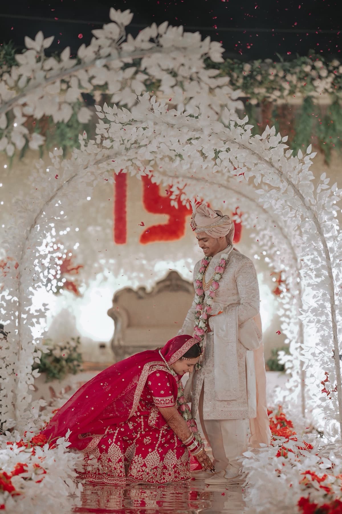 Bride in red lehenga bows to the groom's feet beneath a white floral wedding arch with sparklers
