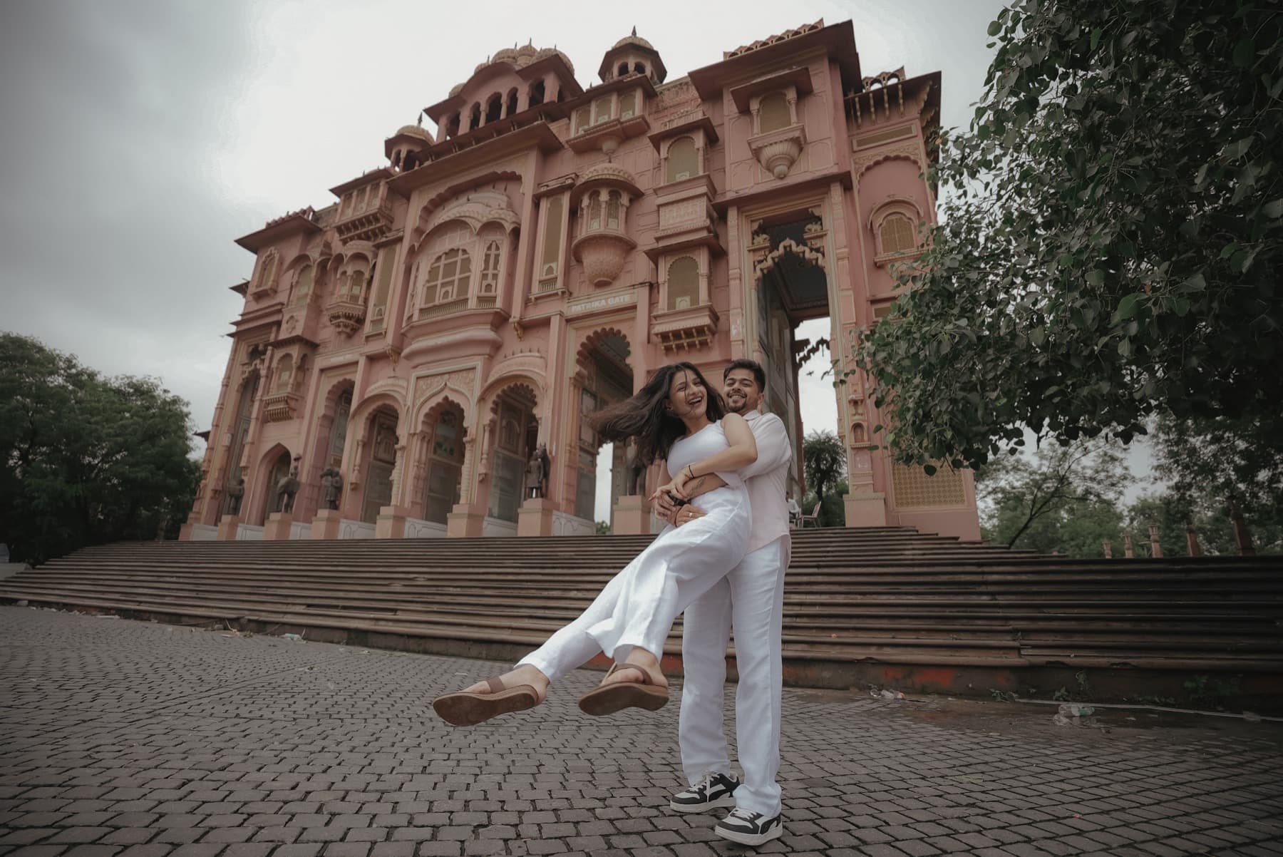 Man lifts and spins a woman in white on a brick plaza before a pink palace facade