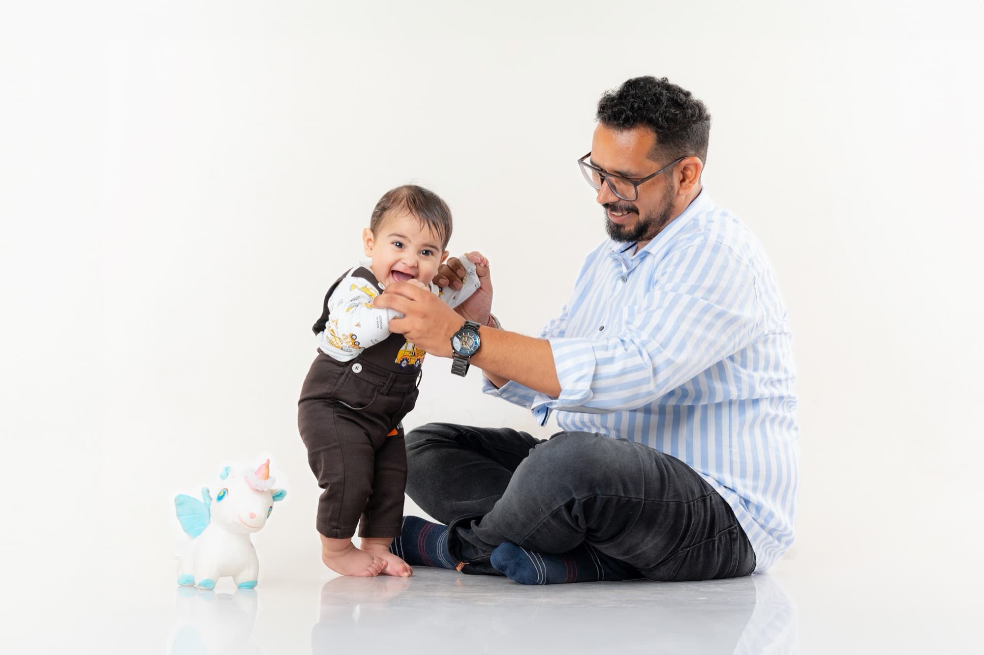Father seated on a white studio floor steadying a laughing toddler on its feet
