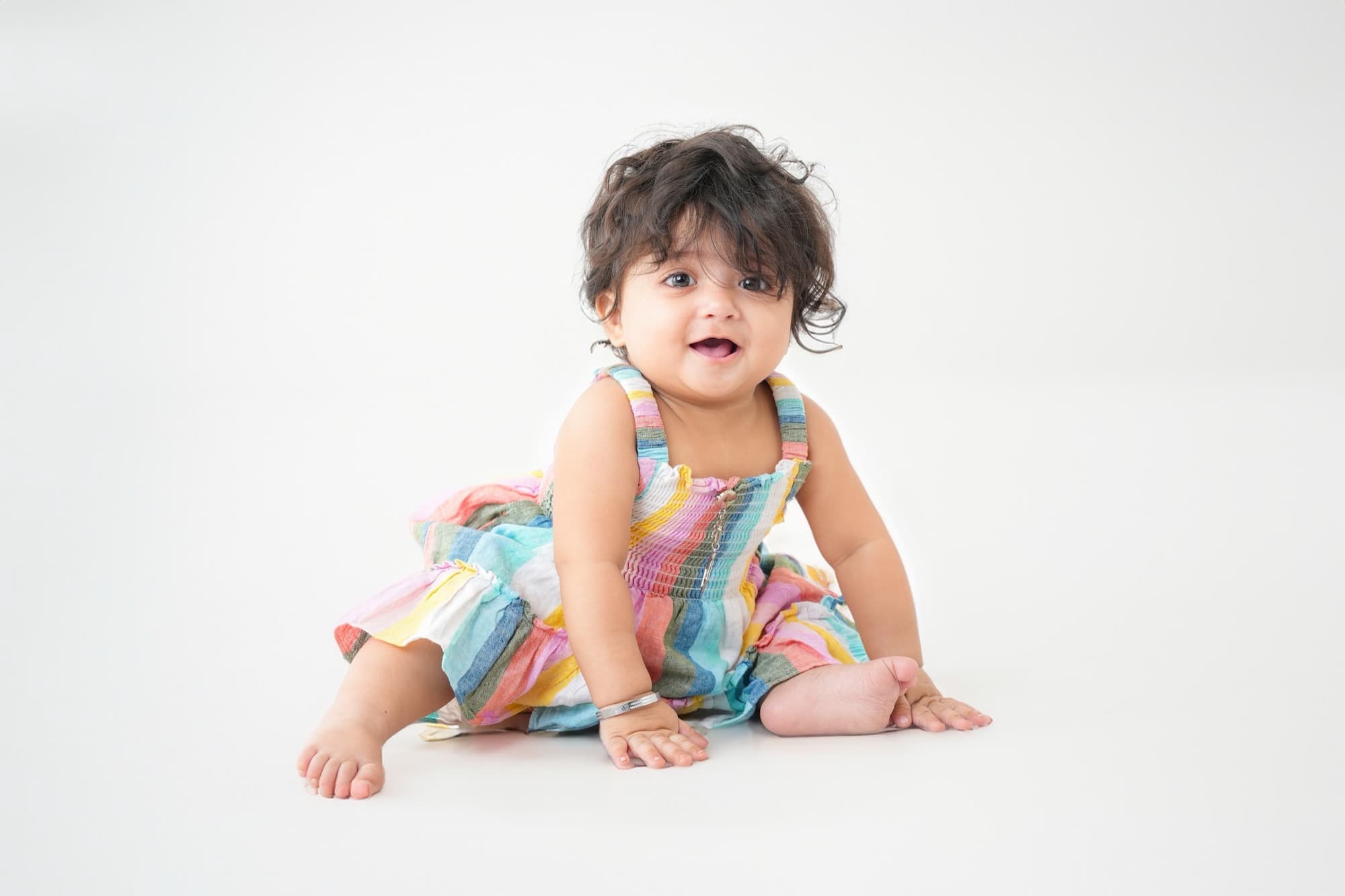 Toddler in a rainbow-striped dress sitting on a white floor, laughing at the camera