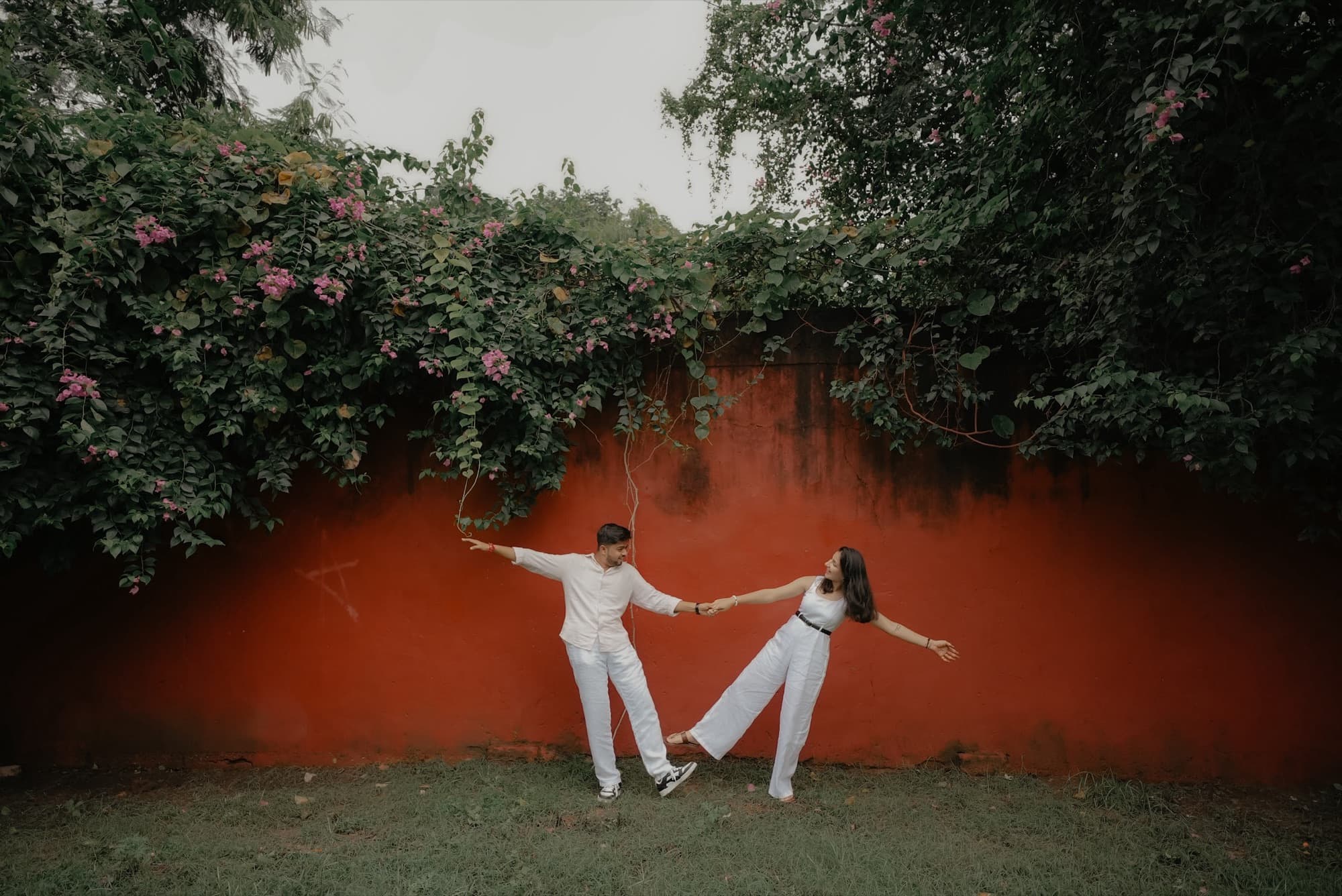Couple in white posing playfully against a red-orange wall draped with bougainvillea