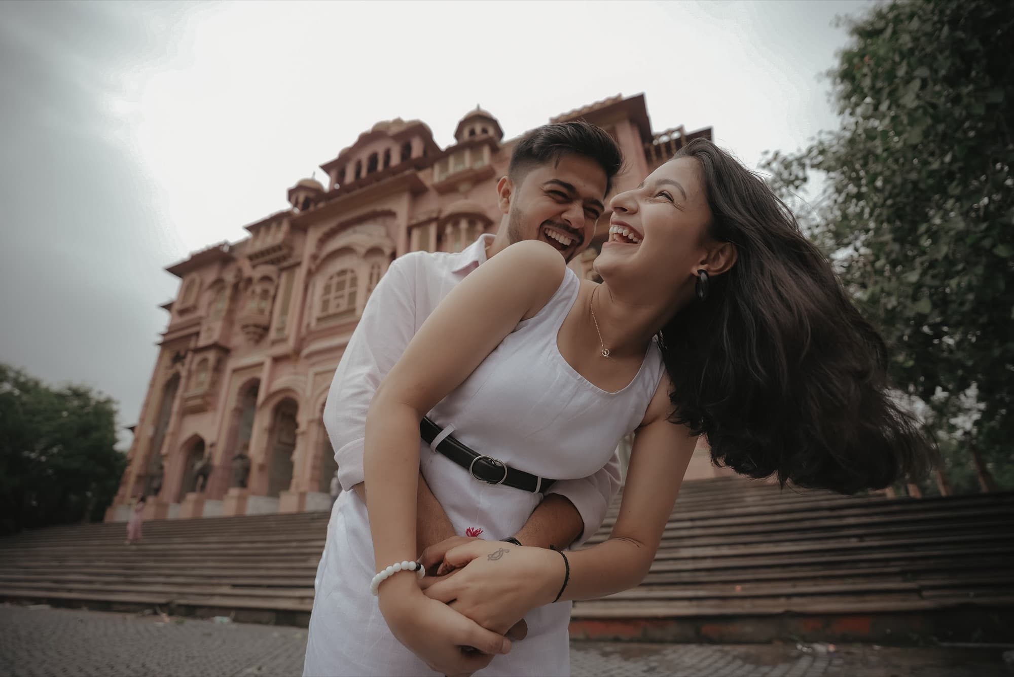 Couple in white laughing together in front of a pink Rajasthani sandstone gateway