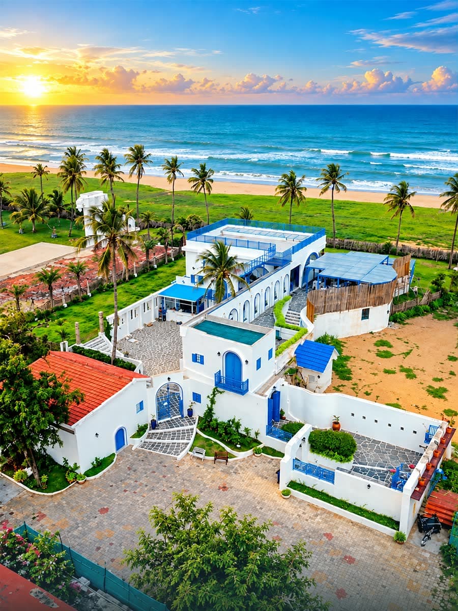 White and blue beachfront cafe with a rooftop terrace, palms and a sunset over the sea behind it, from the air