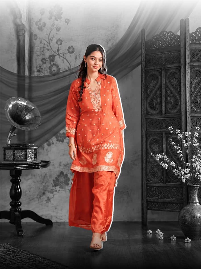 Woman in an orange kurta set with gold embroidery walks towards the camera through a black and white room, a gramophone on a table to her left and a carved wooden screen and white blossoms behind her