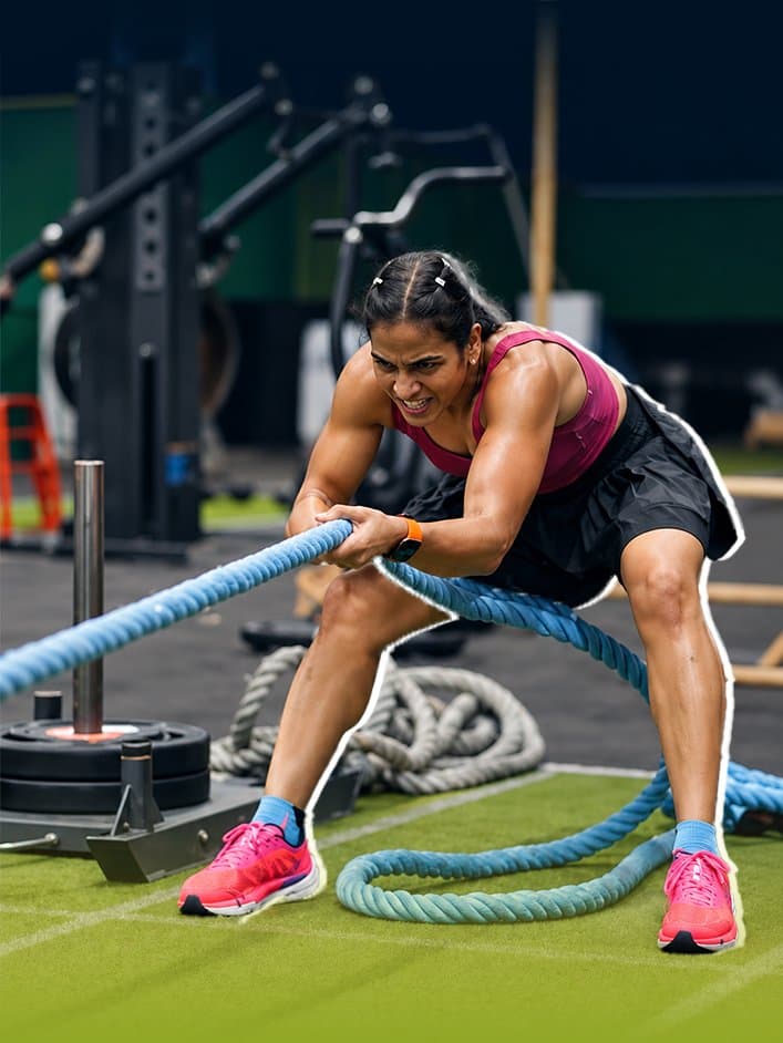 Athlete in a magenta crop top and black shorts grimaces as she whips a pair of battle ropes across the green turf of a gym