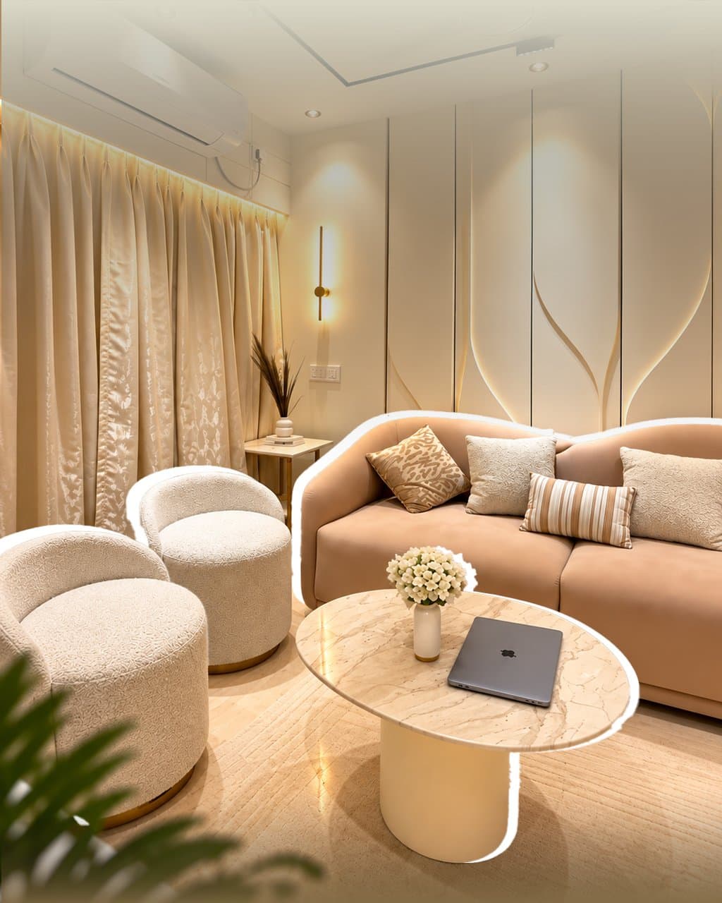 Cream living room with a curved beige sofa, two boucle armchairs and an oval marble coffee table holding a laptop and white flowers, wall panels edged in gold behind