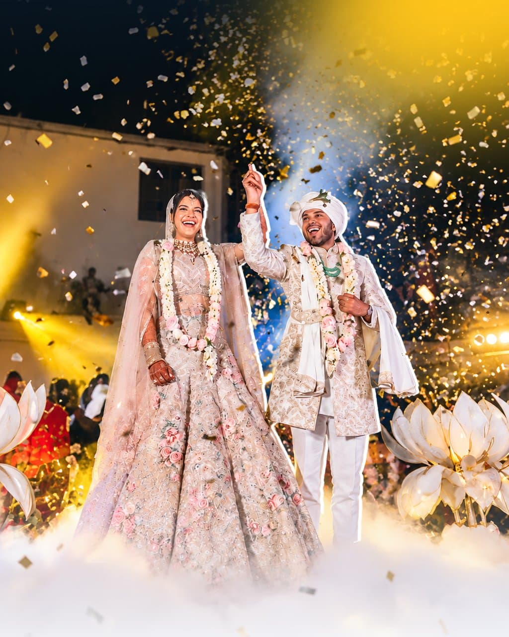 Laughing bride in a blush floral lehenga and groom in a cream sherwani and turban raise their joined hands as gold confetti falls, smoke at their feet and white lotus props either side