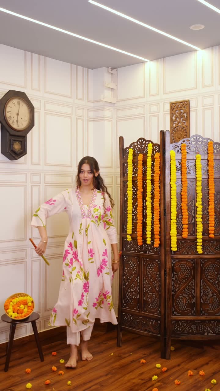 Woman in a white floral anarkali stands barefoot before carved wooden screens hung with marigold strings